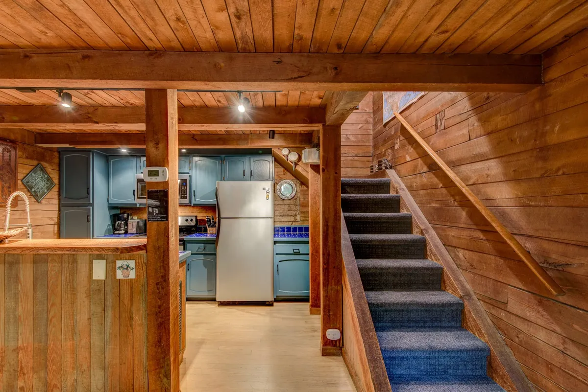 A warm and inviting kitchen area featuring blue cabinets, a wooden countertop, and wooden beams and paneling throughout. The open layout includes a view of the staircase leading to the upper level, giving a sense of connection to the rest of the living space.