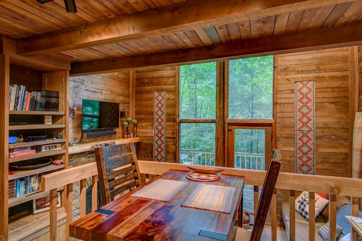 An inviting dining area with a solid wooden table set near large windows that frame lush wilderness views. A curated bookshelf and decorative wall panels add layers of personality and warmth, and the overall setting is both elegant and welcoming, ideal for intimate meals or quiet reading sessions.