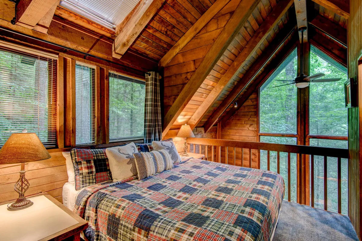 A warmly lit sleeping area featuring plaid bedding, exposed wooden beams, a large window letting in natural light, and a bedside lamp. The view suggests a peaceful atmosphere perfect for relaxation or reading by the window.