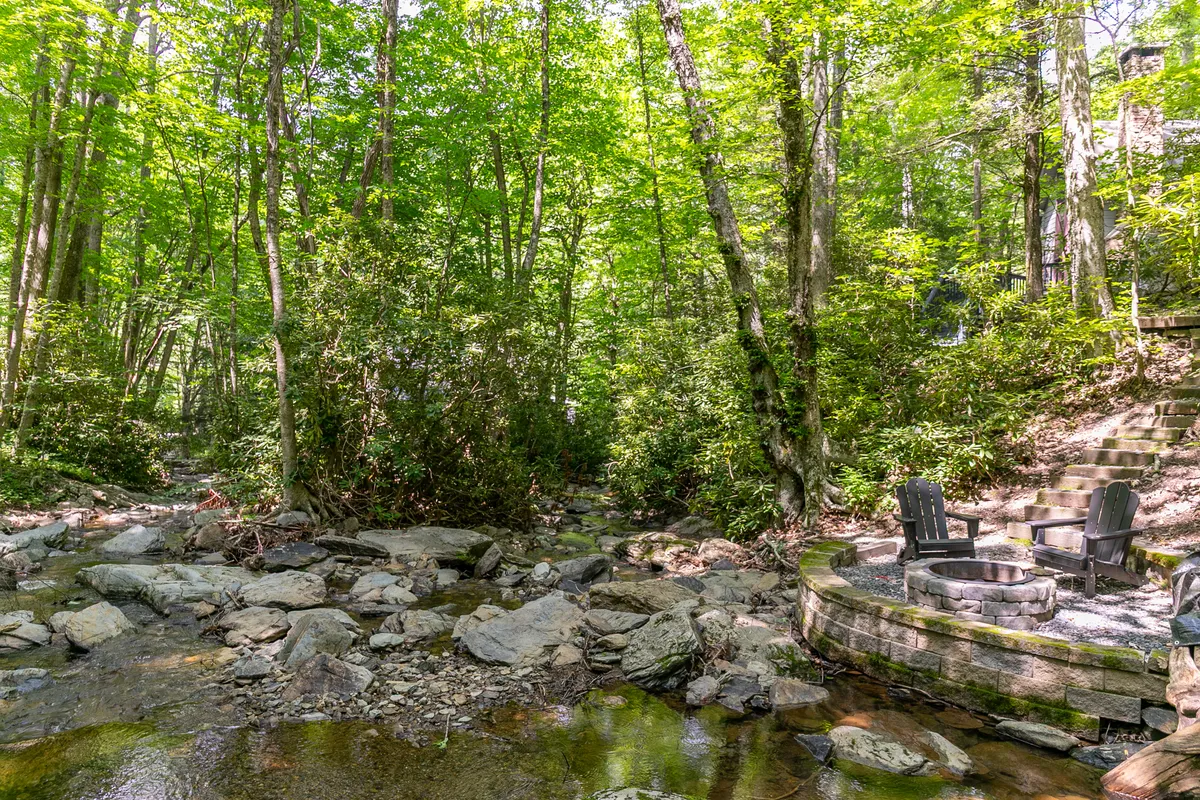 This exterior image shows a tranquil stream winding through lush greenery, with a stone fire pit nestled nearby and surrounded by trees. The scene evokes a sense of peaceful seclusion and immersion in nature, with ample space for outdoor relaxation and exploration.