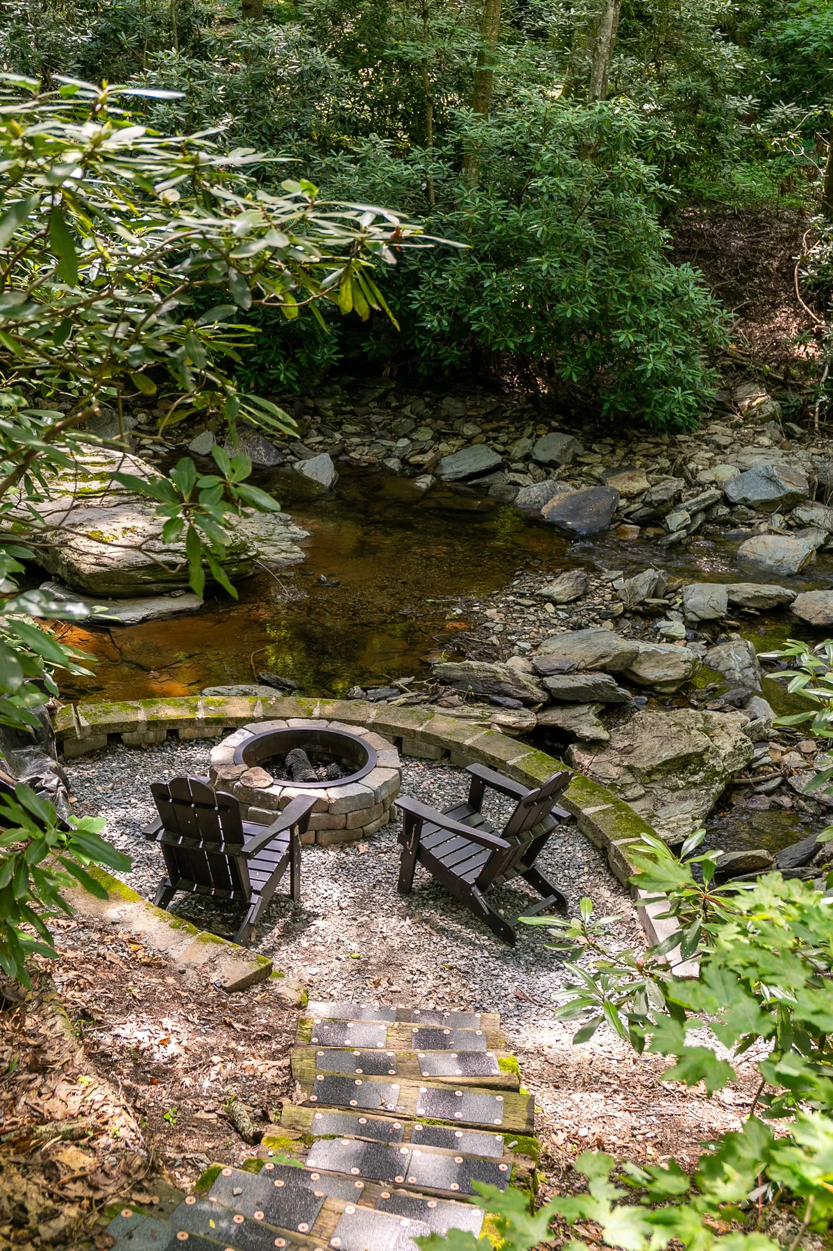 A picturesque scene of a stone fire pit surrounded by Adirondack chairs overlooking the creek and forest. The cozy seating area invites gatherings, storytelling, and relaxation by the fire, set against a lush natural backdrop.