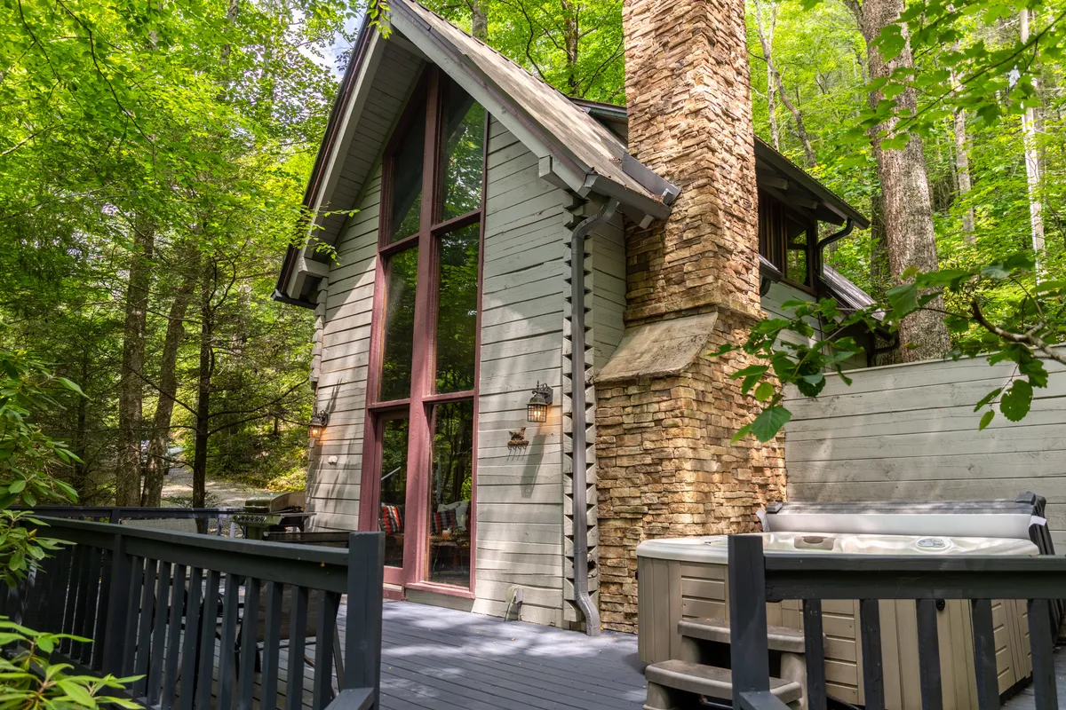 The image showcases the inviting exterior of a cozy mountain cabin nestled in lush woods, featuring tall windows that glow with warm light, a handsome stone chimney, and a spacious wooden deck with a bubbling hot tub. The tranquil creekside setting is perfect for relaxation, with sunset views and a sense of peaceful seclusion.