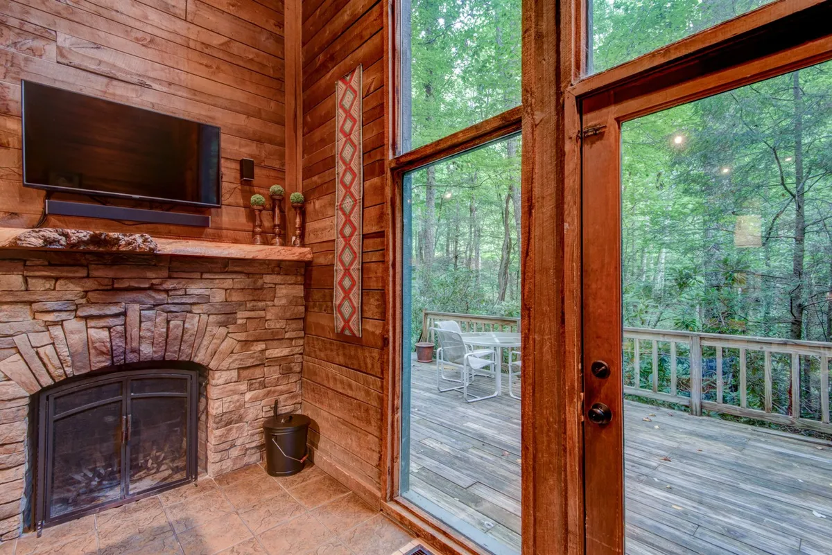 A vibrant living space featuring a stone fireplace as the focal point, a widescreen television, large windows with forest views, and decorative elements that blend rustic and modern aesthetics. The layout encourages relaxation, conversation, and entertainment with friends or family.
