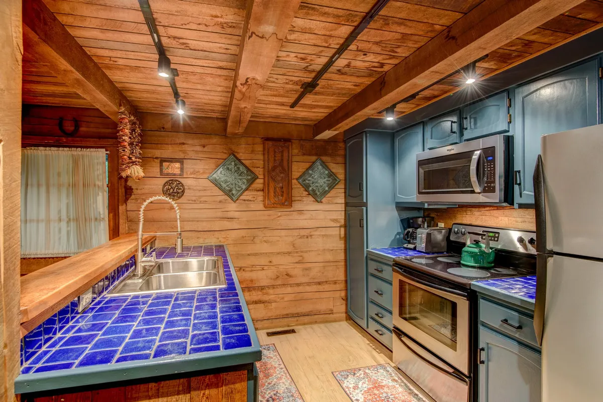 A cozy kitchen scene with a blue tile countertop, wooden beams, modern appliances, and decorative wall elements. The arrangement subtly displays the well-stocked utensils and the artisanal detailing of the space.