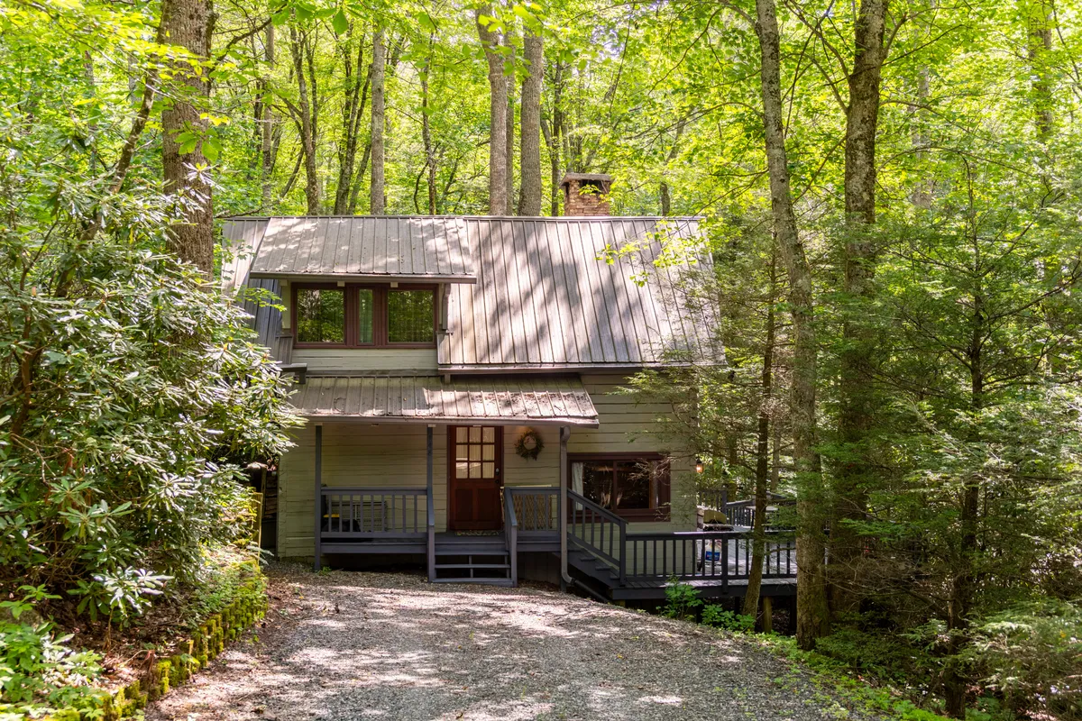 A welcoming view of the cabin's front door, spacious porch, and distinctive roofline set among tall trees. The porch is an inviting spot for morning coffee or evening conversations, with forest views underscoring the connection to the local environment and nearby attractions.
