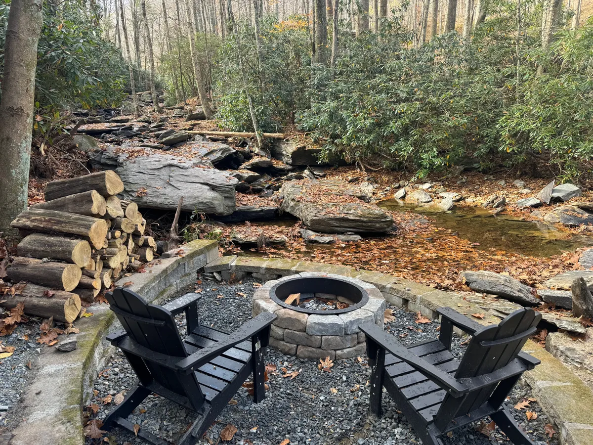 A cozy fire pit area with Adirondack chairs is set beside a gently flowing stream. Stacked firewood and the rustic ambiance invite guests to gather around for storytelling, roasting marshmallows, or stargazing after a day of outdoor excitement.