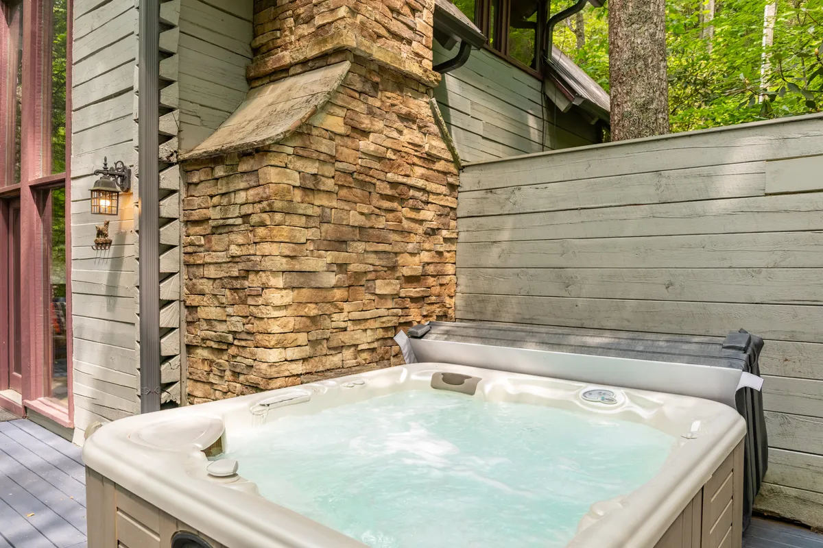The exterior of the cabin featuring a bubbling hot tub set against a wooden wall and stone chimney. Surrounded by natural greenery, the scene invites guests to unwind outdoors, whether stargazing at night or enjoying a peaceful morning soak.