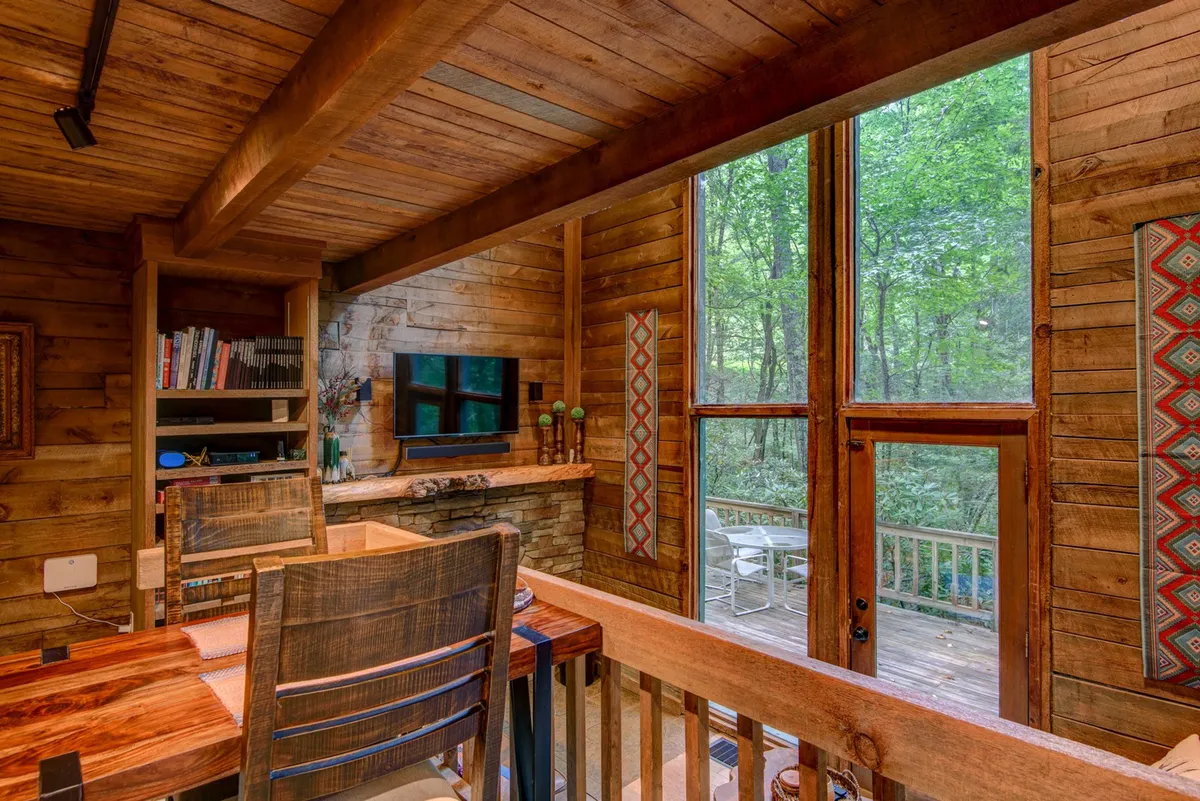 A tranquil living space featuring a stone fireplace, large windows with forest views, a wooden desk, and tasteful decor. The cozy setup invites relaxation by the fire on winter evenings or enjoying morning coffee while gazing at the natural surroundings.