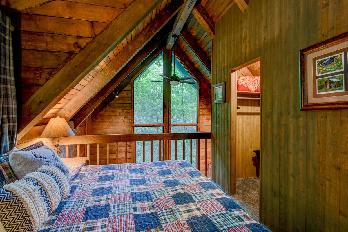 A cozy bed dressed in a patterned quilt sits beneath wooden beams and large windows. The windows frame green, leafy views, and a warm lamp on the bedside table adds a comforting glow. The space exudes tranquility and warmth, making it an inviting spot to unwind or enjoy a private retreat.
