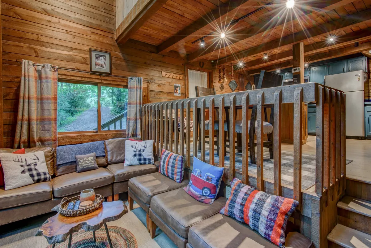 A warm, inviting living space with a comfortable sectional sofa, decorative pillows, wooden railings, and wide windows letting in natural light. The arrangement is clearly designed for family togetherness, offering space for games, meals, and relaxed conversation, with glimpses of the outdoors beyond the glass.