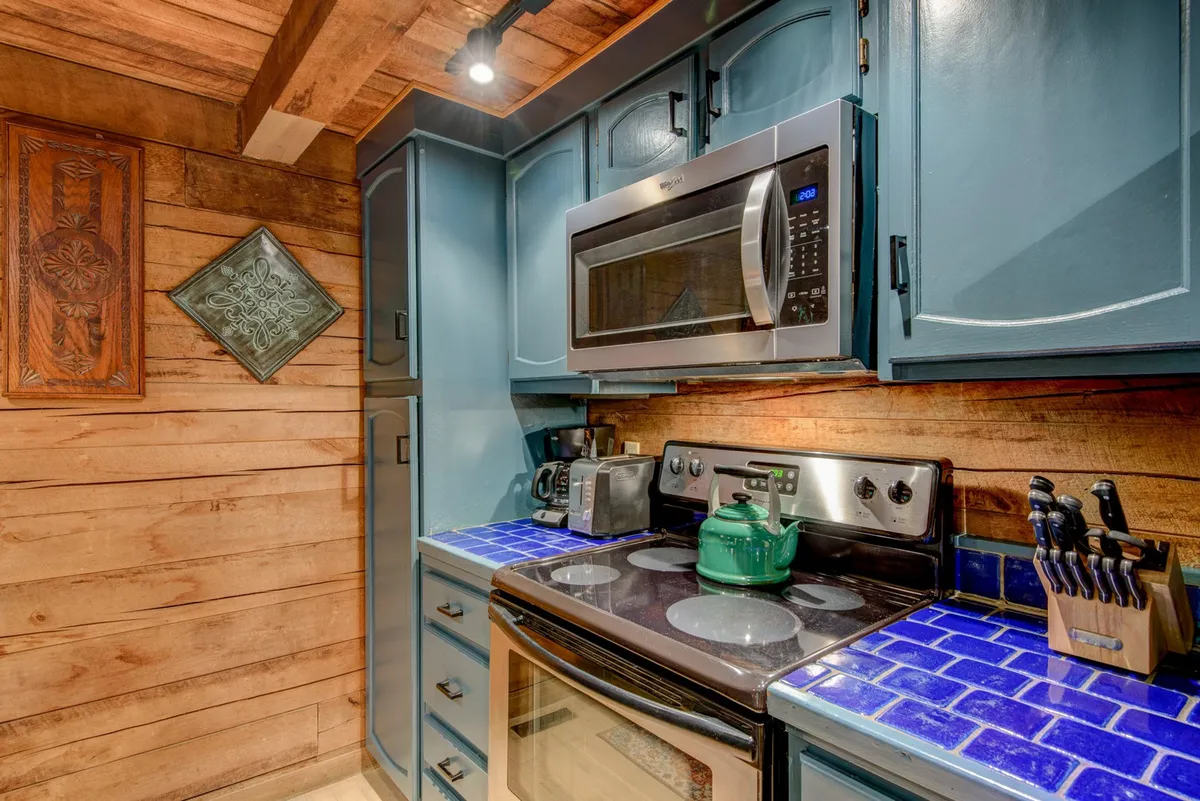 A close-up of the kitchen workspace, featuring blue cabinets, stainless steel appliances, a blue tiled countertop, and a green kettle. The setup illustrates a space ready for meal prep, baking, or gathering with loved ones.